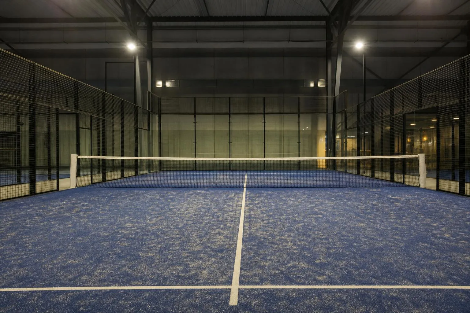 Un court de padel vide, parois vitrées et grillage, filet tendu sur un sol synthétique bleu